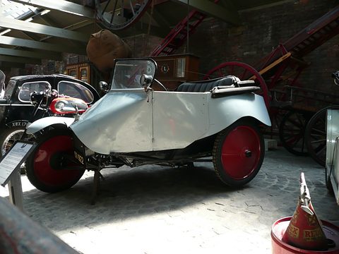 A grey open three-wheeler with red disc wheels, a single front wheel and a windscreen, among motorcycles under a brick shed. Opens a larger view