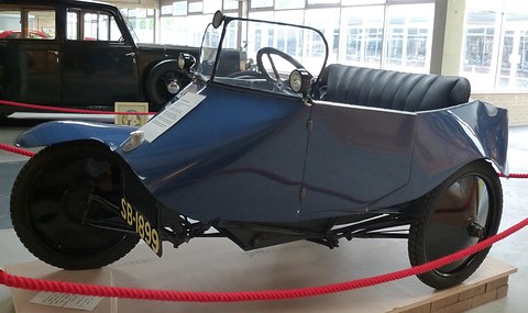 A blue open three-wheeler with a single front wheel under a cycle wing and two lamps, behind a red rope.