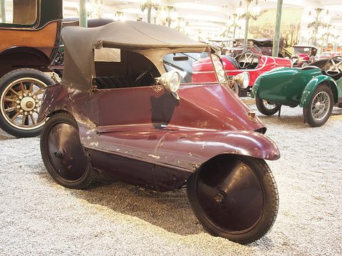 A maroon three-wheeler with faded paint and a black folding hood, standing on gravel among veteran cars. Opens a larger view