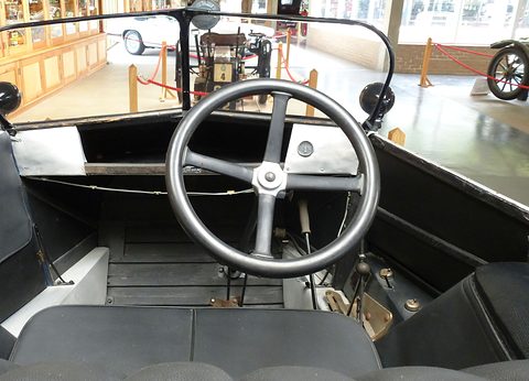 Looking down into a bare cockpit: a four-spoke steering wheel, a black bench seat and a plain wooden floor. Opens a larger view