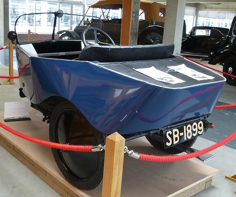 Rear three-quarter view of a blue three-wheeler with a pointed tail, a number plate and two closely set rear wheels. Opens a larger view