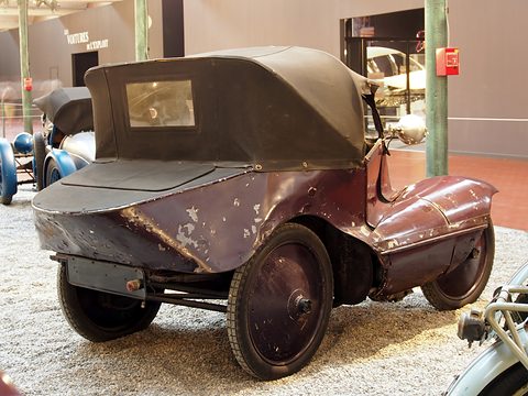Rear three-quarter view of a battered maroon three-wheeler with a black hood and a flat rear deck. Opens a larger view