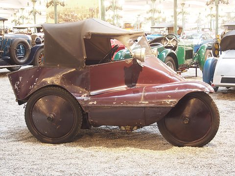 Side view of a maroon three-wheeler with chipped paint, disc wheels and a black hood, on a gravel floor. Opens a larger view