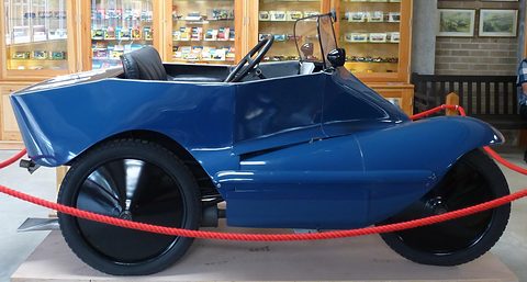 Side view of a long blue boat-shaped body slung low between a front wheel and a rear wheel, cabinets of toy cars behind. Opens a larger view