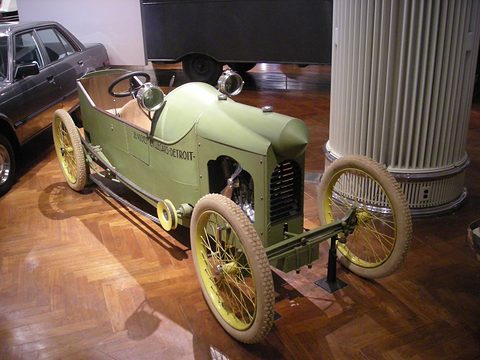 A narrow olive-green tandem cyclecar with a pointed bonnet, louvred nose and yellow wire wheels on a parquet floor.