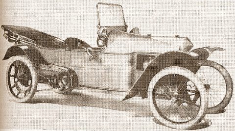 A sepia view of a long, low open cyclecar with wire wheels, a tall windscreen and a spare wheel at its side.