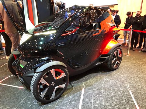 Black and orange narrow two-seat concept with a door raised on a patterned floor, visitors behind a rope. Opens a larger view