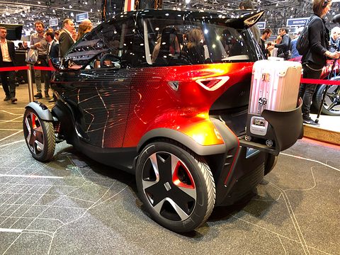 Rear of a black and orange narrow concept with a scooter-style rear case and large wheels, visitors behind. Opens a larger view