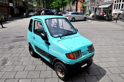 Pale turquoise boxy microcar with amica lettering on the nose, standing on paving in a sunlit street with parked cars. Opens a larger view