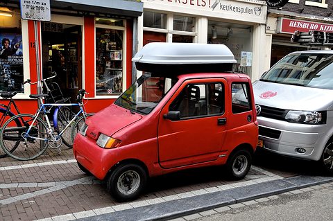 Small rounded red two-door microcar with a grey roof box, parked on a brick pavement beside bicycles and a white van. Opens a larger view