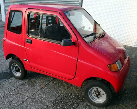Red rounded two-door microcar seen from the front right, parked on paving before a white tiled wall. Opens a larger view