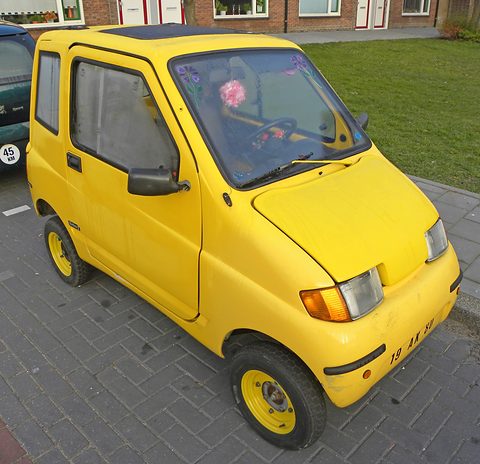 Rounded yellow two-door microcar with yellow wheels and a flower decoration on the dashboard, parked on brick paving. Opens a larger view