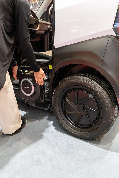A person wheeling a black battery case with a white ring out from beside a car’s rear wheel.