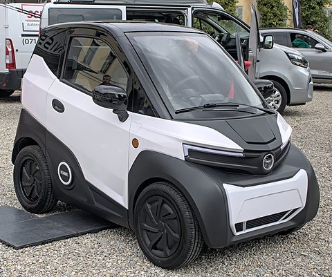 White and black two-seat microcar with a bonnet mesh and round wheel-arch badge on gravel, vans behind. Opens a larger view