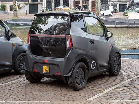 Rear of a grey two-seat microcar with a dark tailgate and yellow plate on brick paving beside a canal. Opens a larger view