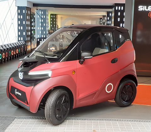 Red two-seat microcar with black roof and front, its bonnet open, inside a showroom with city graphics. Opens a larger view