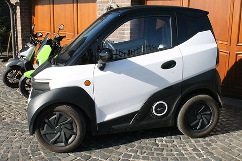 Side view of a white and black two-seat microcar on cobbles in front of wooden garage doors and scooters. Opens a larger view