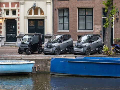 Three small grey and black microcars parked in a row by canal houses, boats moored in front. Opens a larger view