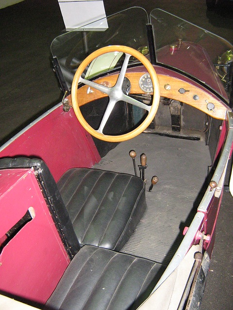 A wooden steering wheel and wooden dash over two black leather seats and a gear lever in a red body. Opens a larger view