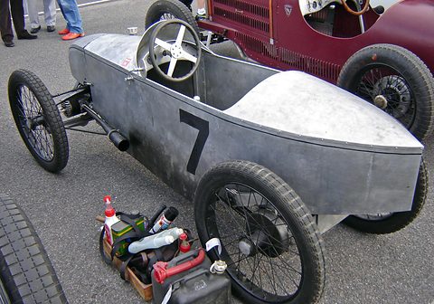 A small unpainted racing cyclecar with number 7 on its flank, a tool box and bottles on the ground beside it. Opens a larger view