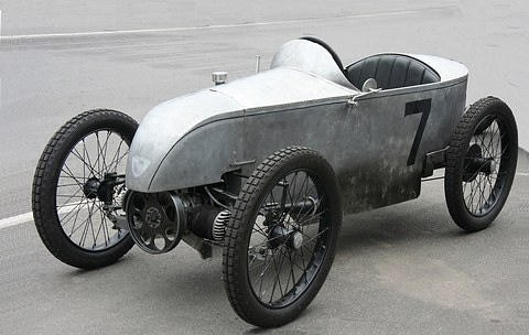 A tiny unpainted racing cyclecar with wire wheels, a pointed tail and an engine jutting out beside the nose. Opens a larger view