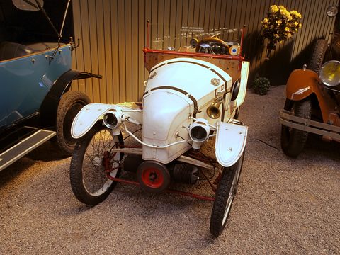 Front view of a white cyclecar with two horn-like cylinders sticking out each side of the nose and a red flywheel. Opens a larger view