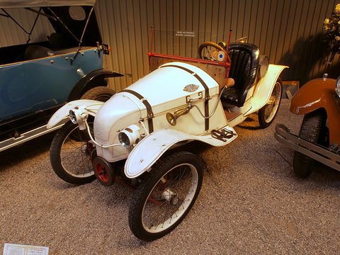 A white two-seat cyclecar with leather bonnet straps, cycle wings and red wheels on a gravel floor. Opens a larger view