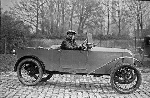 A man in a cap and leather jacket at the wheel of a small open two-seater with wire wheels on cobbles.