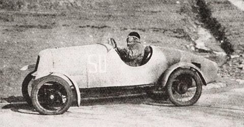 A half-tone of a small pale racing car with a pointed nose and disc wheels, its driver in a helmet waving. Opens a larger view