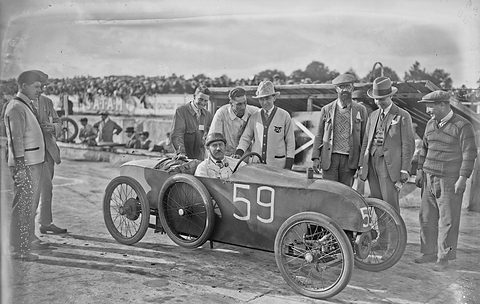 A driver sits in a narrow racing cyclecar numbered 59 while men in caps and hats stand around it in the paddock. Opens a larger view