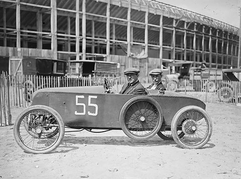 Two men sit side by side in a low, slab-sided racing cyclecar with wire wheels, a grandstand under construction behind. Opens a larger view