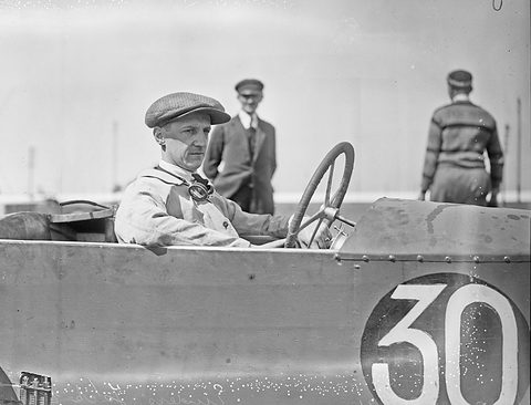 A man in a flat cap sits at the wheel of a low racing car with number 30 on its flank.