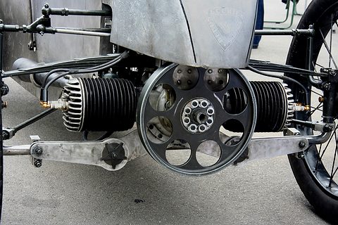 Head-on view of a flat-twin engine with finned barrels either side of a large drilled flywheel under an aluminium nose. Opens a larger view