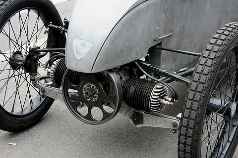 Two finned cylinders stick out sideways from a perforated flywheel at the front of a small car, between wire wheels.