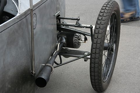 Close view of a wire front wheel, a sprung axle, an exhaust pipe and a finned cylinder beside an aluminium body. Opens a larger view