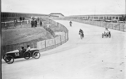 A low cyclecar rounds a curve of a concrete track beside a fence, two motorcyclists and another small car further on.