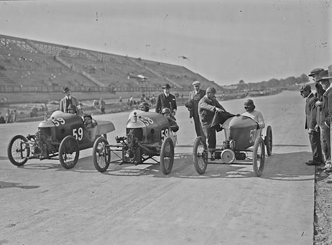 Three small racing cyclecars side by side on a banked concrete track, drivers in helmets, officials in hats beside them. Opens a larger view