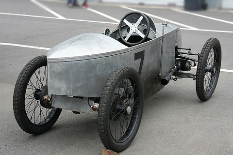 Rear three-quarter view of a narrow unpainted cyclecar with a pointed tail, a steering wheel and wire wheels. Opens a larger view