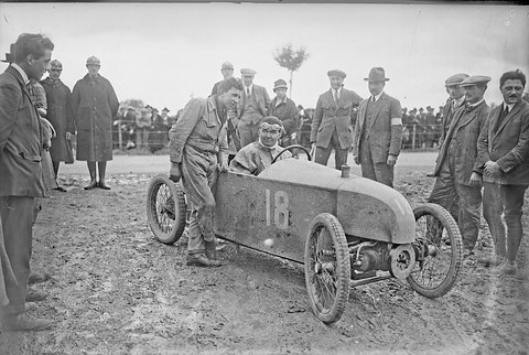 A mud-spattered small pointed racing car numbered 18, its helmeted driver seated, men in caps and suits round it. Opens a larger view