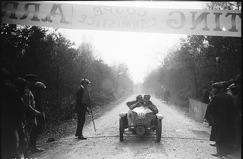 A small racing car with two men aboard drives down a misty woodland road under a banner. Opens a larger view