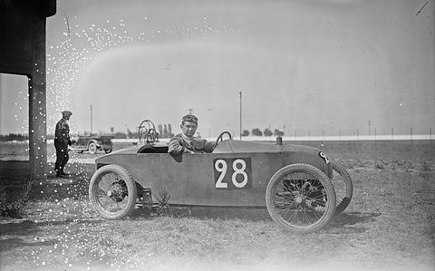 A narrow racing cyclecar with number 28 and a seated driver on rough grass, a man standing by a pillar at the left. Opens a larger view