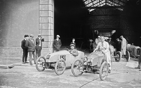 Two small pale racing cyclecars with drivers aboard before a large garage doorway, men in caps standing by. Opens a larger view
