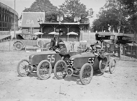 Two small racing cyclecars with exposed front engines and wire wheels parked together, crews aboard, flags on a pavilion behind. Opens a larger view