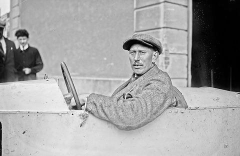 A man in a flat cap and tweed coat looks back over his shoulder from the cockpit of a pale racing car. Opens a larger view
