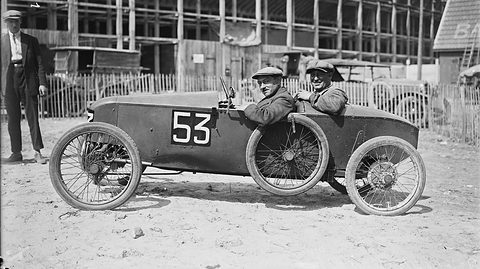Two smiling men in caps in a low racing cyclecar numbered 53, a man in a suit standing beside it on sandy ground.