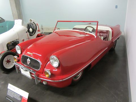 Low red open two-seater with an oval mesh grille, round headlamps and a chrome-rimmed windscreen, on a dark museum floor.