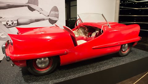 Long low red open two-seater with tail fins and a red steering wheel, on a black plinth before a mural of aircraft. Opens a larger view