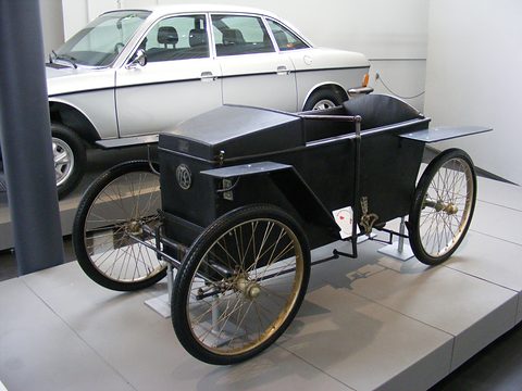 A small black box-bodied electric car with tall wire wheels on a pale plinth, a silver saloon behind.