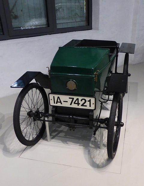 Front view of a narrow green box-bodied electric car with wire wheels and a white number plate. Opens a larger view