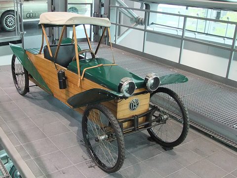 A slatted varnished-wood car with green wings and seats, a cream canvas hood and spoked wheels on a grey tiled floor. Opens a larger view
